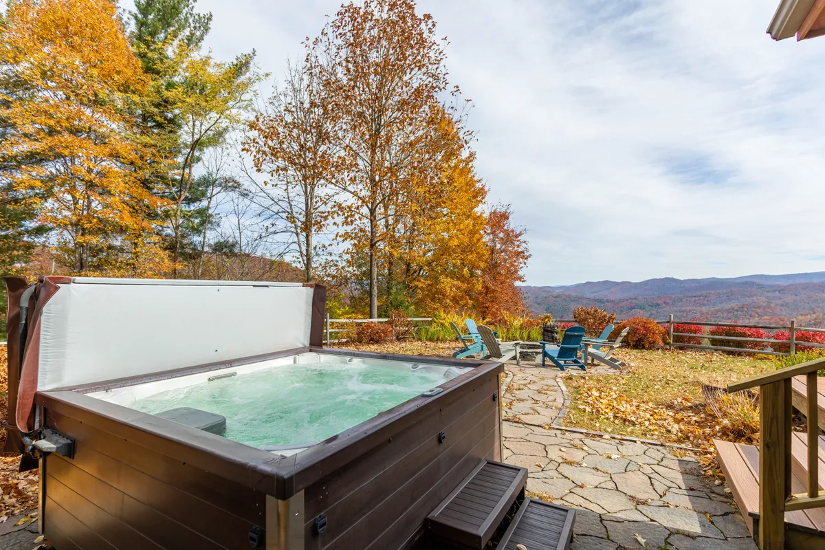 The hero image showcases a stunning mountain lodge surrounded by vibrant autumn foliage. The stone patio features a bubbling hot tub positioned to take in sweeping mountain views, with the warm glow of sunset painting the landscape. The serene scene invites guests to relax in the hot tub or gather with friends on the patio, all while taking in the tranquil, colorful beauty of the Blue Ridge Mountains.