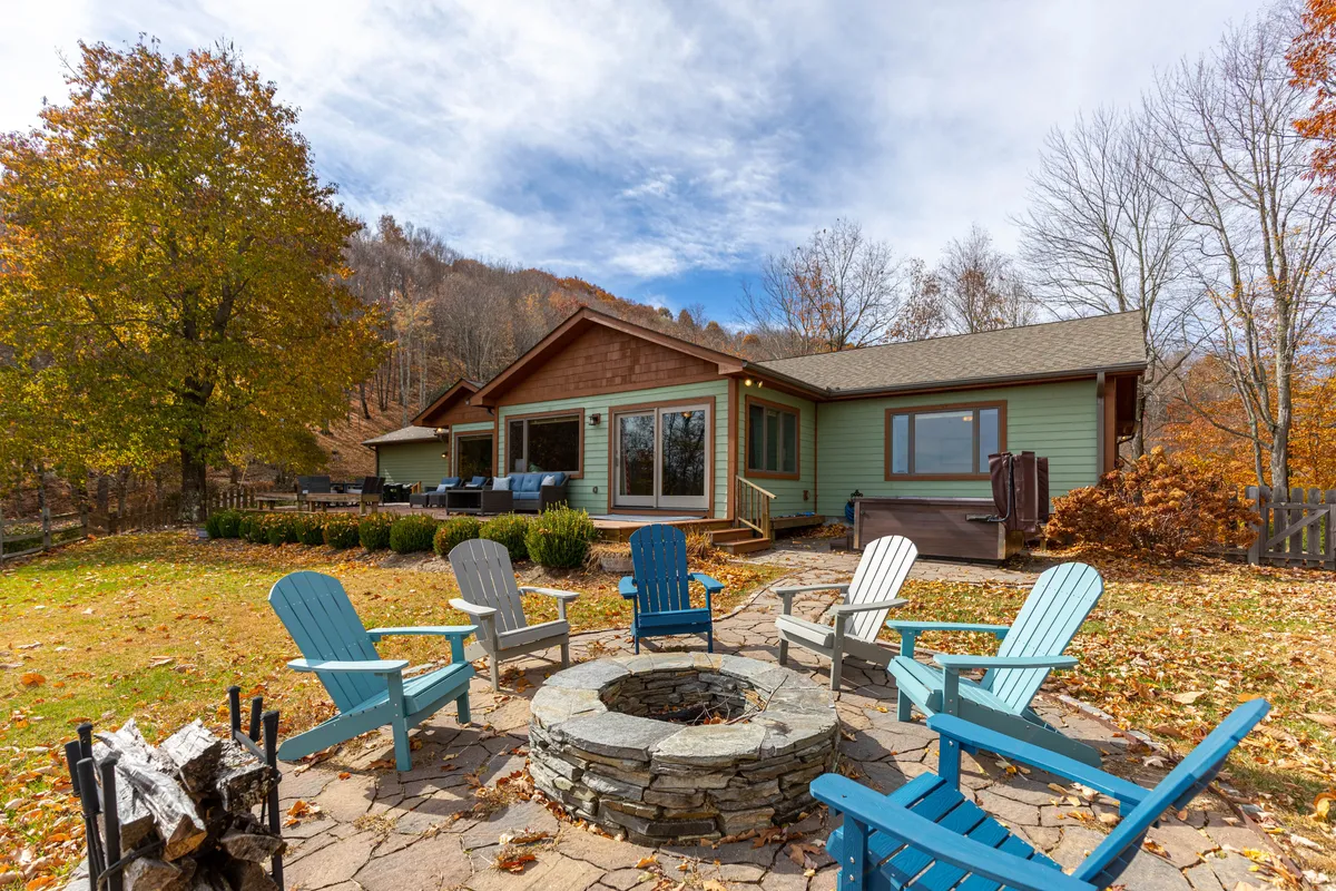 This image captures the inviting exterior fire pit area with Adirondack chairs arranged around a glowing fire. The house is visible in the background, framed by natural surroundings, suggesting a perfect setting for group gatherings, evening relaxation, and enjoying mountain views.