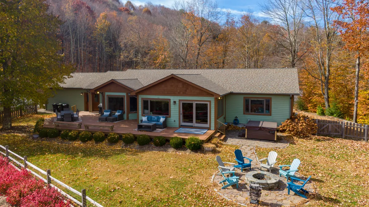 A wide shot of the modern house exterior, showcasing the generous deck, fire pit area with Adirondack chairs, and autumnal trees. Guests are seen gathering around the fire pit, with the sunset casting a warm glow over the scene.