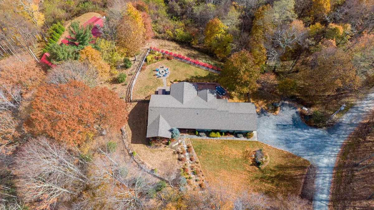A wide shot of the property showcases the sprawling home, beautiful landscaping, a fire pit area, and a winding driveway. The spacious yard and colorful trees create a welcoming setting for outdoor family gatherings, picnics, and play.