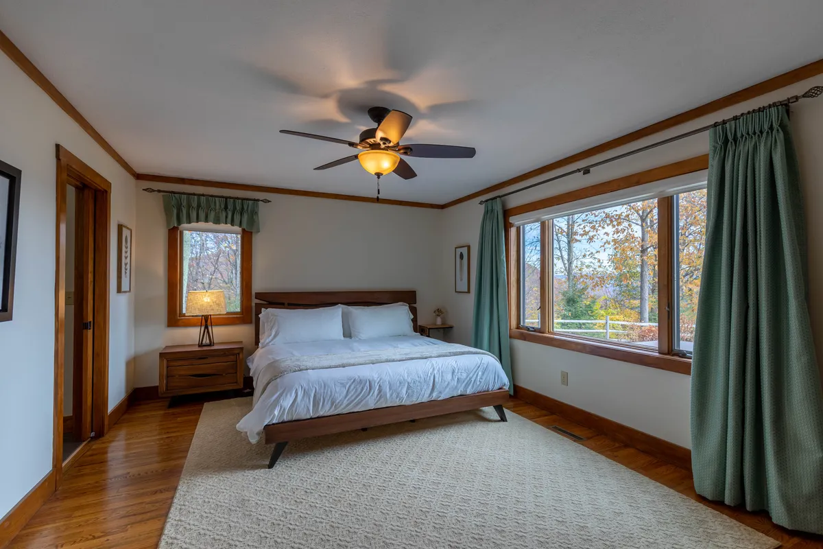 This image features a serene king bedroom with a plush bed, large windows letting in natural light, a nightstand, and a lamp. The calming palette and uncluttered decor emphasize tranquility and comfort, echoing the restful nature of the suite.