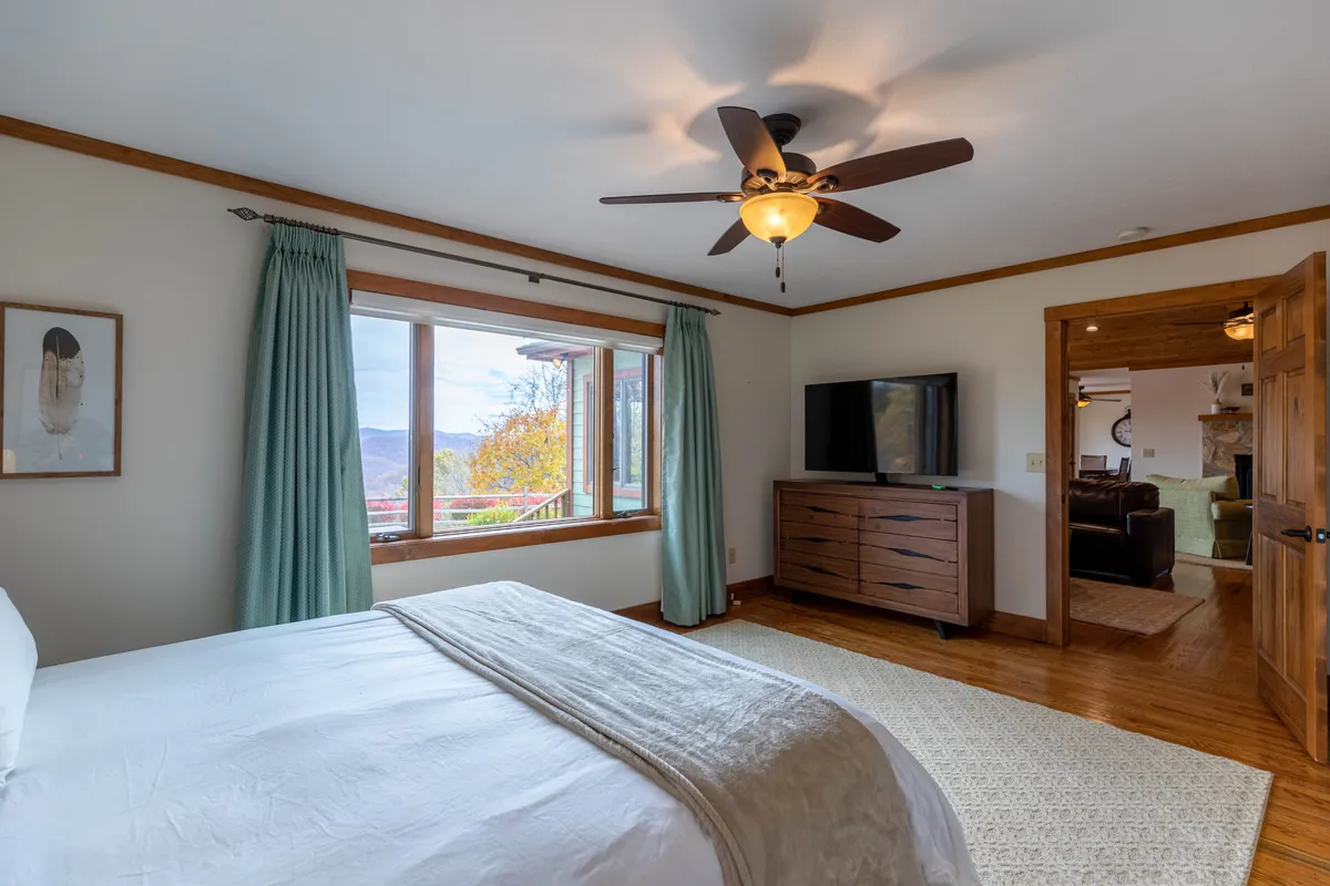 A spacious king bedroom with a comfortable bed positioned to take advantage of scenic views through a large window. The wooden dresser with TV and premium linens highlight both luxury and relaxation.