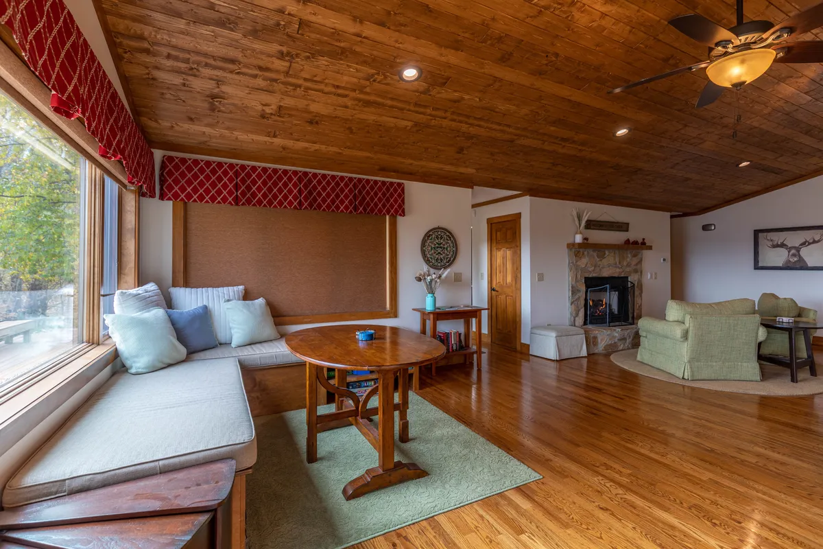 A bright, welcoming living area with expansive windows offering glimpses of the surrounding landscape. The room features a cozy seating area, a wooden table perfect for morning coffee, and a fireplace – all oriented to take advantage of the natural light and outdoor views.