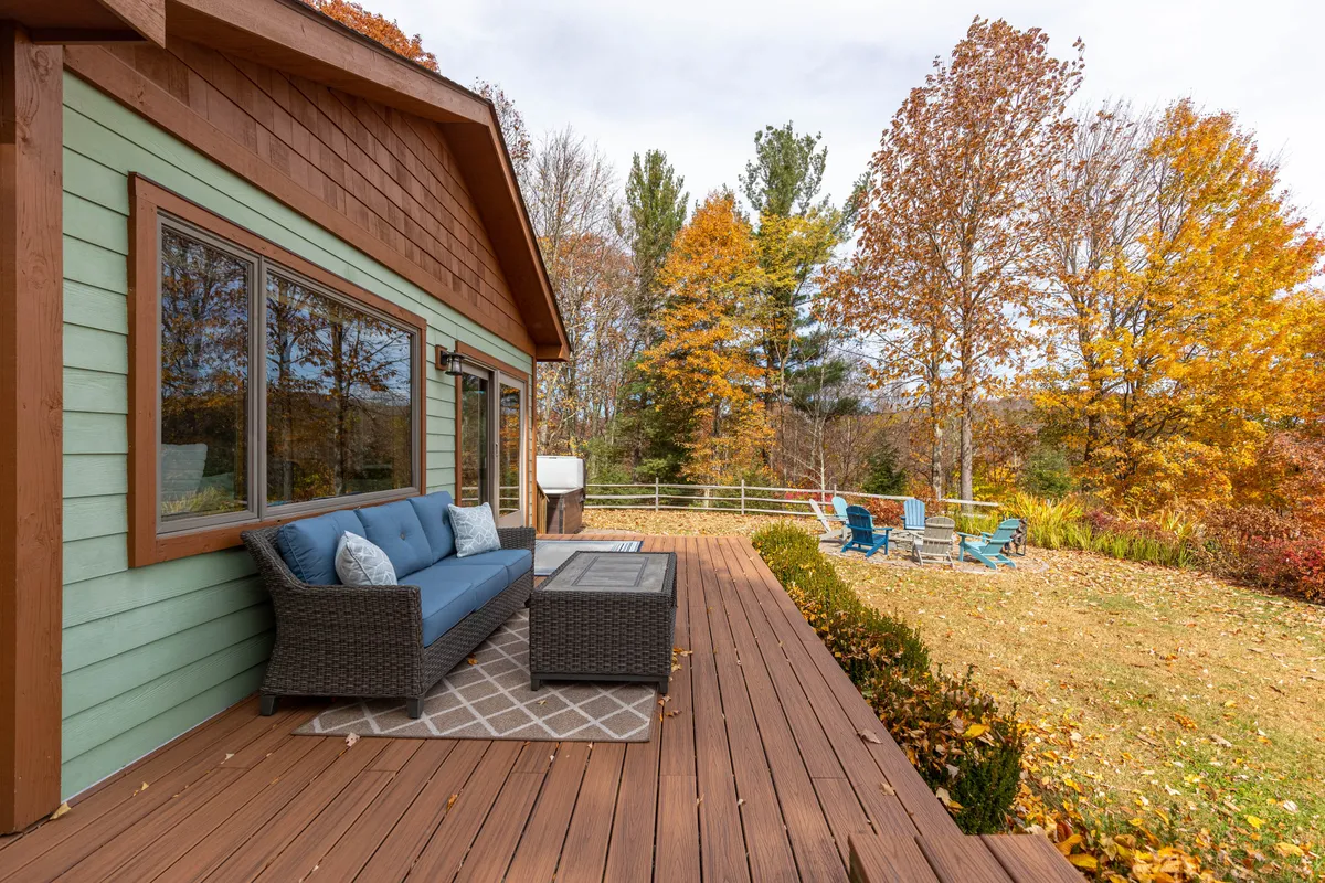 A cozy blue outdoor sofa sits on a wooden deck surrounded by vibrant autumn foliage. Patio chairs provide additional seating, and the fenced feeling of the space suggests safety for pets. The inviting atmosphere encourages both relaxing with loved ones and sharing the outdoors with four-legged companions.