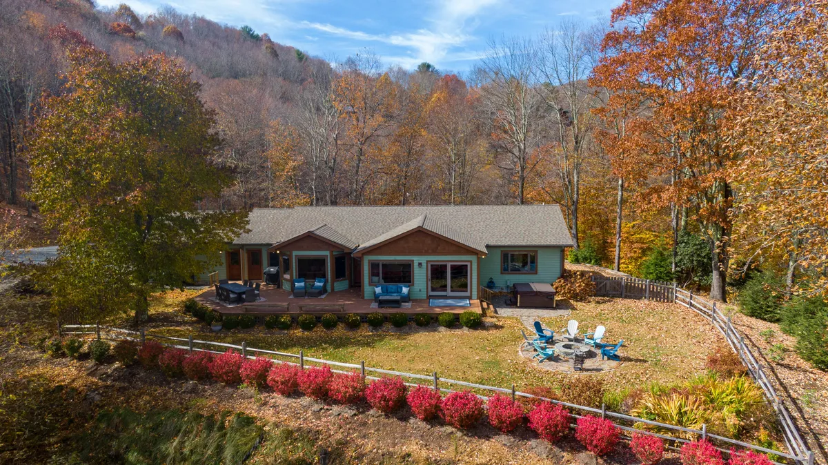 The house is framed by lush, colorful foliage, with a welcoming outdoor seating area and a fire pit in the foreground. The space is perfect for group gatherings, play, or cozy evenings around a bonfire, and the open yard suggests plenty of room for pets to explore safely under supervision.
